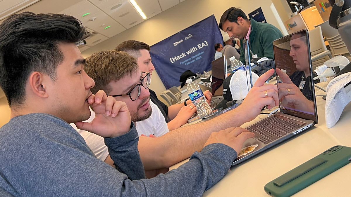 In a large conference room full of people in the background, the photos focuses on four people stare intently at their computer screens with one person pointing at their teammate’s screen.