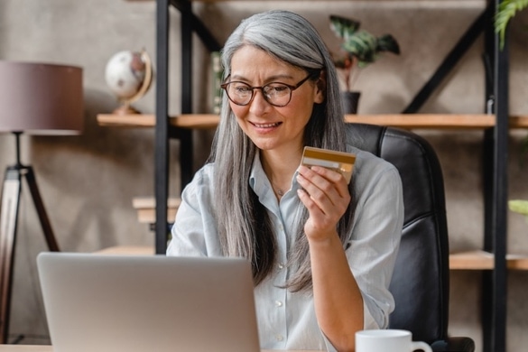 A person sitting in front of a bookcase at a desk looking at a laptop while holding a credit card in their hand