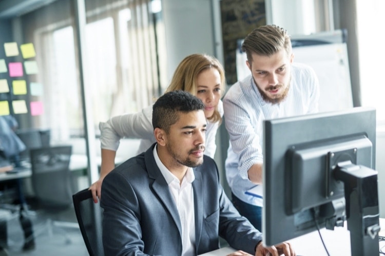 Two people standing with one person sitting at a desk looking at a computer monitor in an office