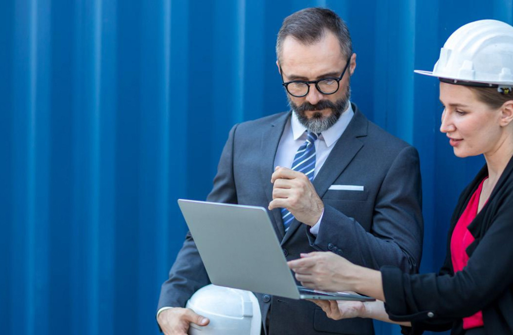 Man wearing a business suit standing next to a woman in a suit and they are looking at a tablet device.